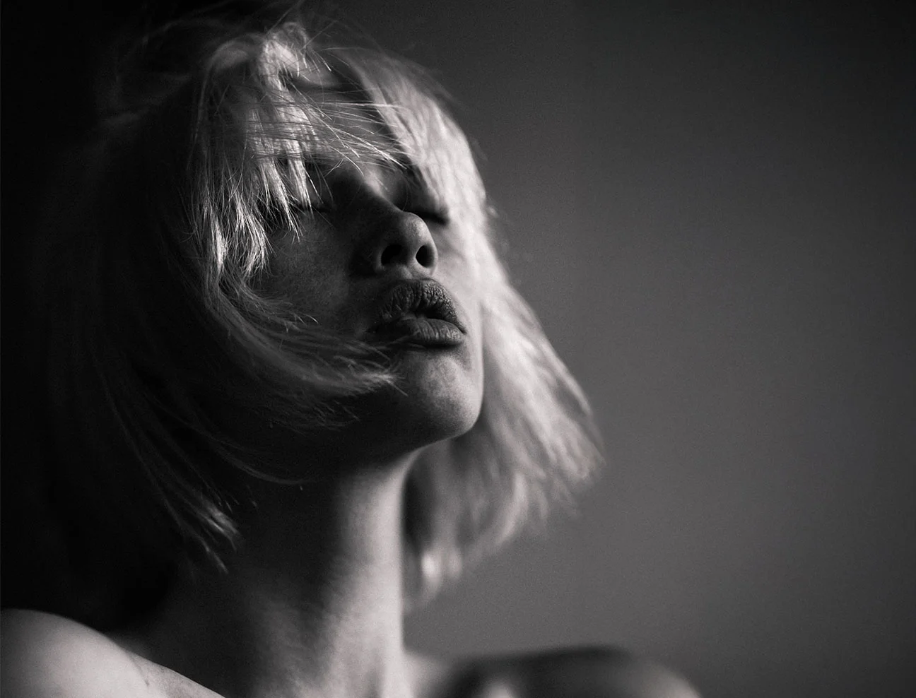 A dramatic black-and-white, close-up portrait of a woman with short hair and eyes closed, tilting her head back as if catching a breeze.