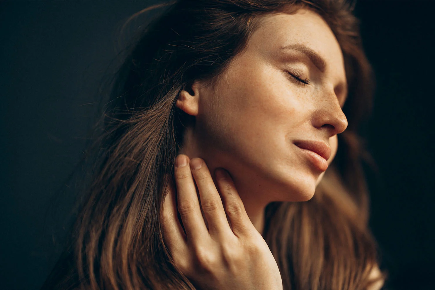 A close-up profile of a woman with freckles and long brown hair. She has her eyes closed and a peaceful expression, gently touching her neck with one hand against a dark, moody background.