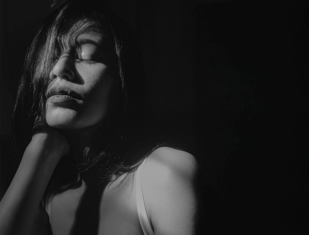 A low-light, black-and-white artistic shot of a woman with her eyes closed and head tilted back. Deep shadows create a dramatic, atmospheric feel.