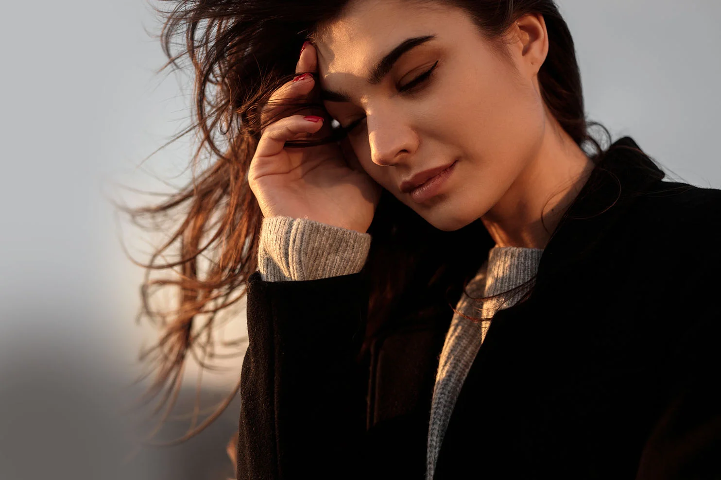 A woman with dark hair and red nail polish rests her hand on her forehead, eyes closed in the warm glow of sunset. She is wearing a black coat over a gray sweater.