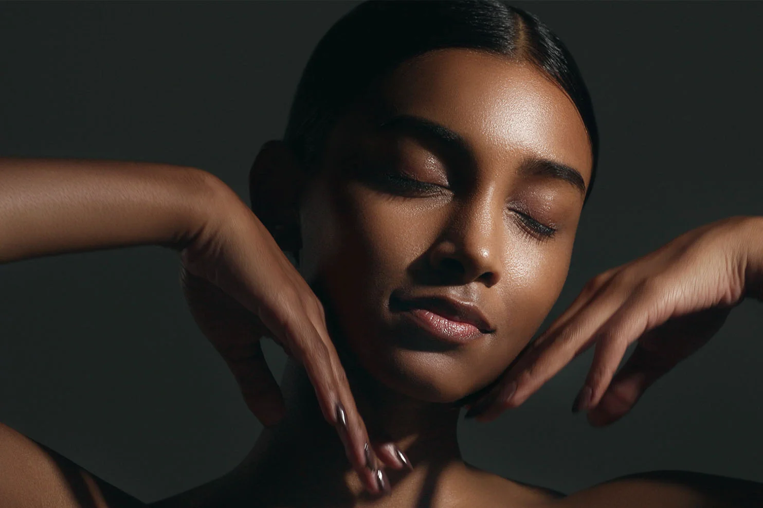 A close-up of a woman with deep skin tones and slicked-back hair. She has her eyes closed and her hands framing her face, with high-contrast lighting emphasizing the glow of her skin.