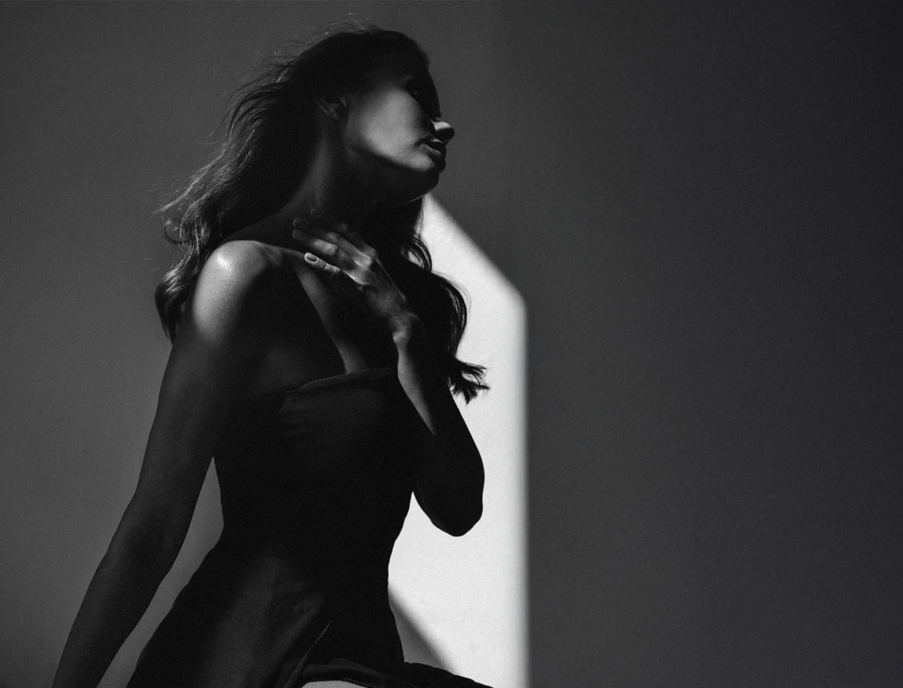 A black-and-white silhouette-style shot of a woman in a dark dress. She is turned in profile, touching her neck as a sliver of light illuminates her shoulder and face.
