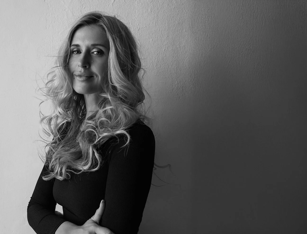 A black-and-white portrait of a woman with long, curly blonde hair. she stands with her arms crossed against a plain wall, smiling gently at the camera.