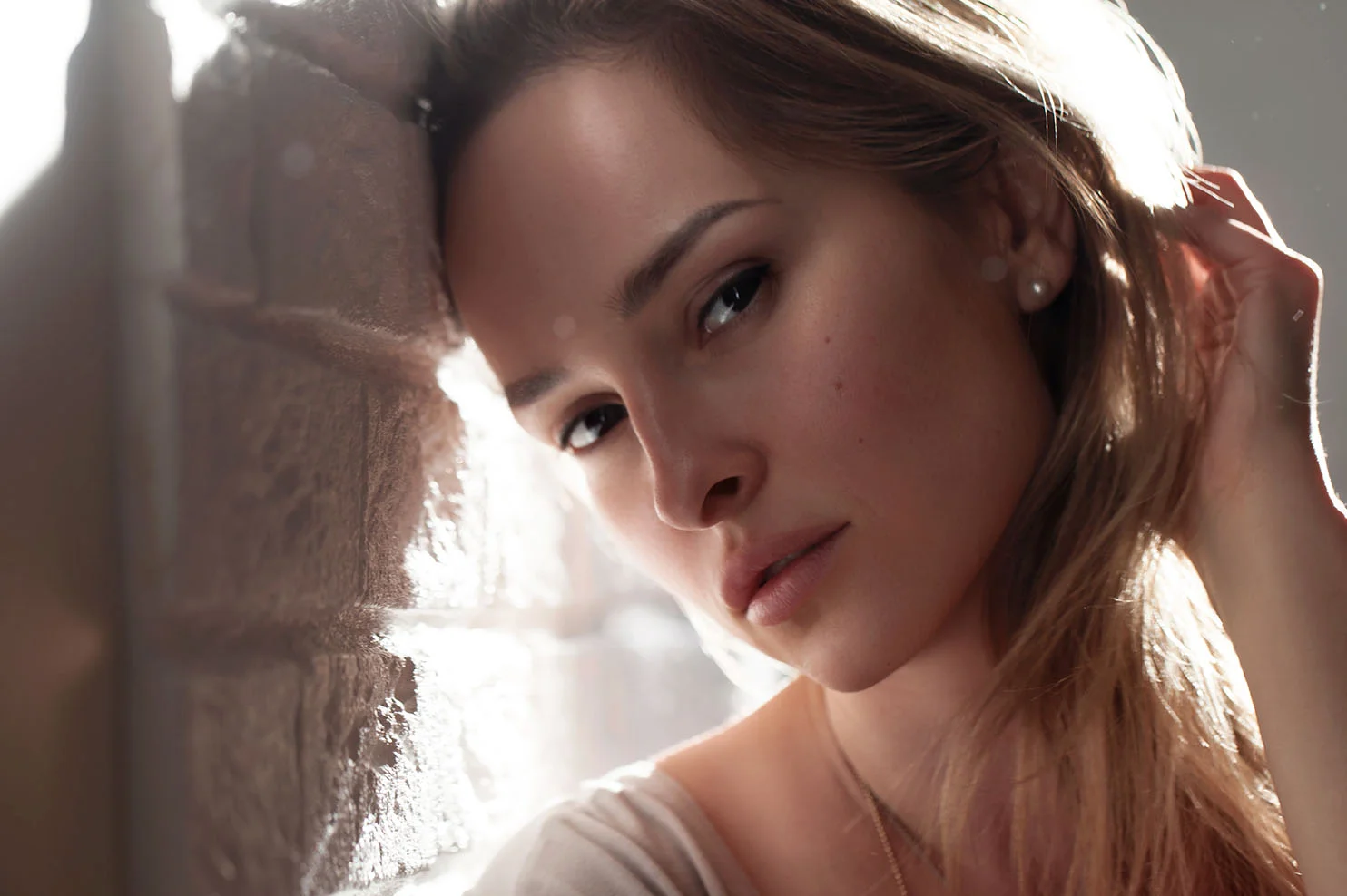 A woman with blonde hair leans her head against a white brick wall. Bright sunlight from behind creates a soft lens flare and highlights the texture of her hair and skin.