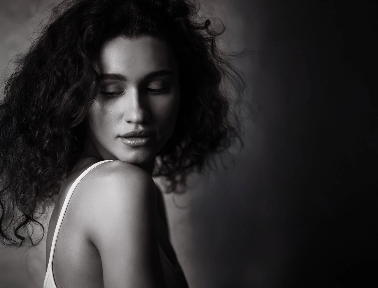 A woman with long curly hair looks over her shoulder with her eyes downcast. The lighting creates a soft, moody atmosphere.