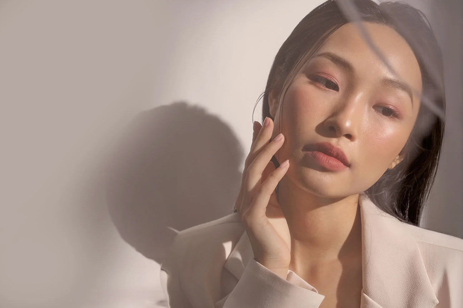 A woman with dark hair and elegant makeup poses with her hand near her face. Harsh, artistic lighting creates a dramatic shadow on the wall behind her, highlighting the contours of her cheekbones.