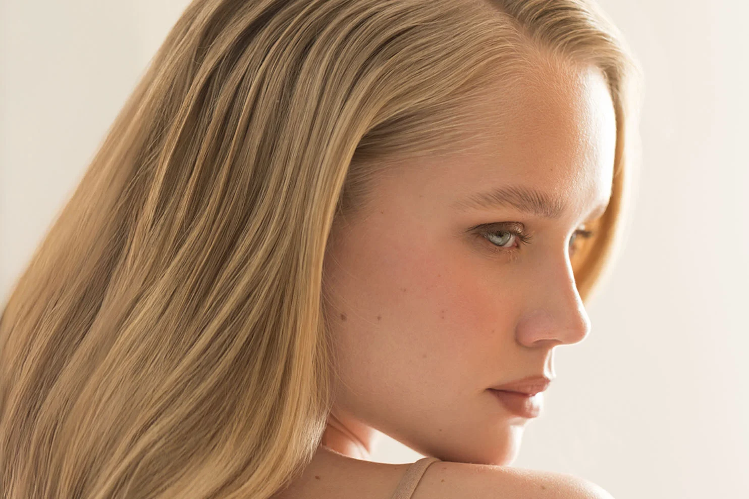 A close-up, side-angle view of a woman with light blonde hair. She is looking over her shoulder with a neutral expression, showcasing a soft, radiant complexion.