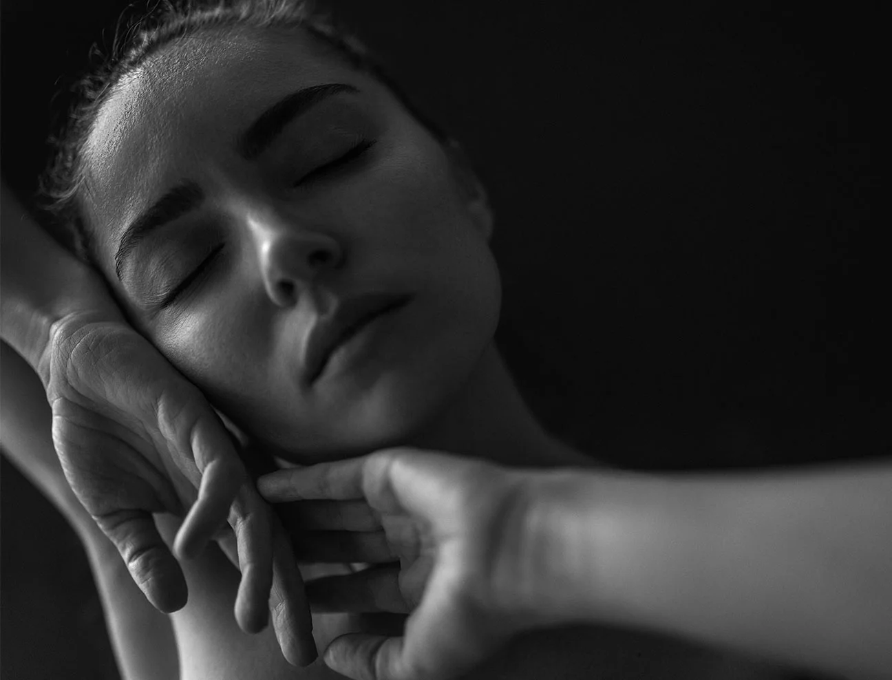 A black and white, close-up artistic shot of a woman’s face with her eyes closed. Her hands are gently framing her jawline and neck, emphasizing a sense of calm and skin texture.
