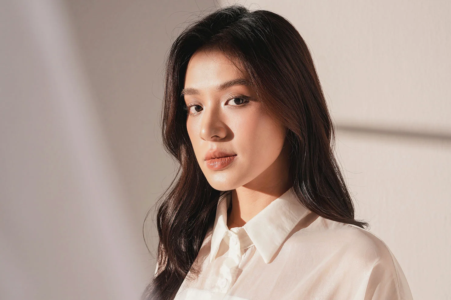 A front-facing portrait of a woman with long dark hair, wearing a white button-down shirt. Warm, natural light and soft shadows from a window fall across her face, emphasizing a smooth and balanced complexion.