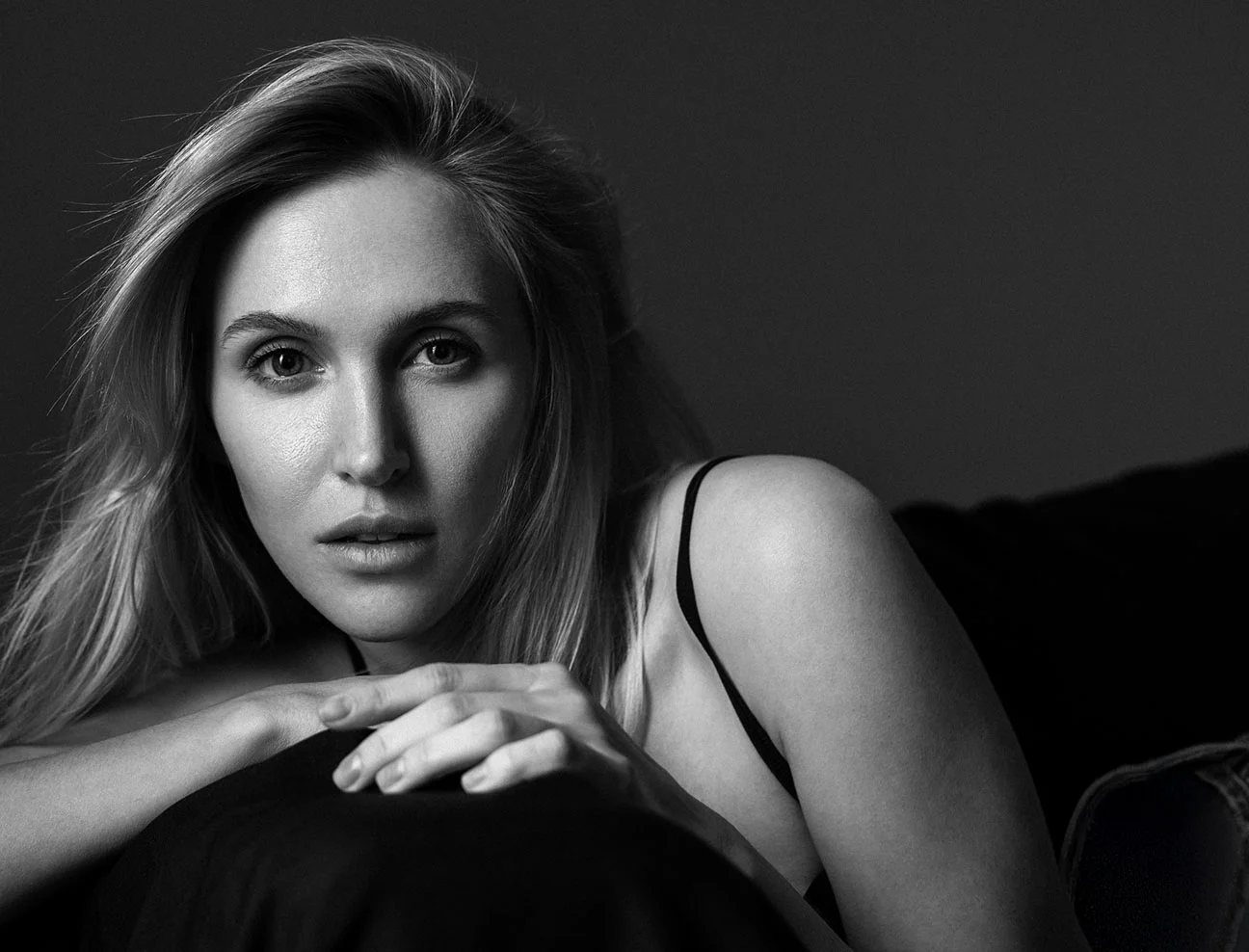 A black and white portrait of a blonde woman leaning forward, resting her chin on her hands. She is looking directly at the camera with a soft, neutral expression.