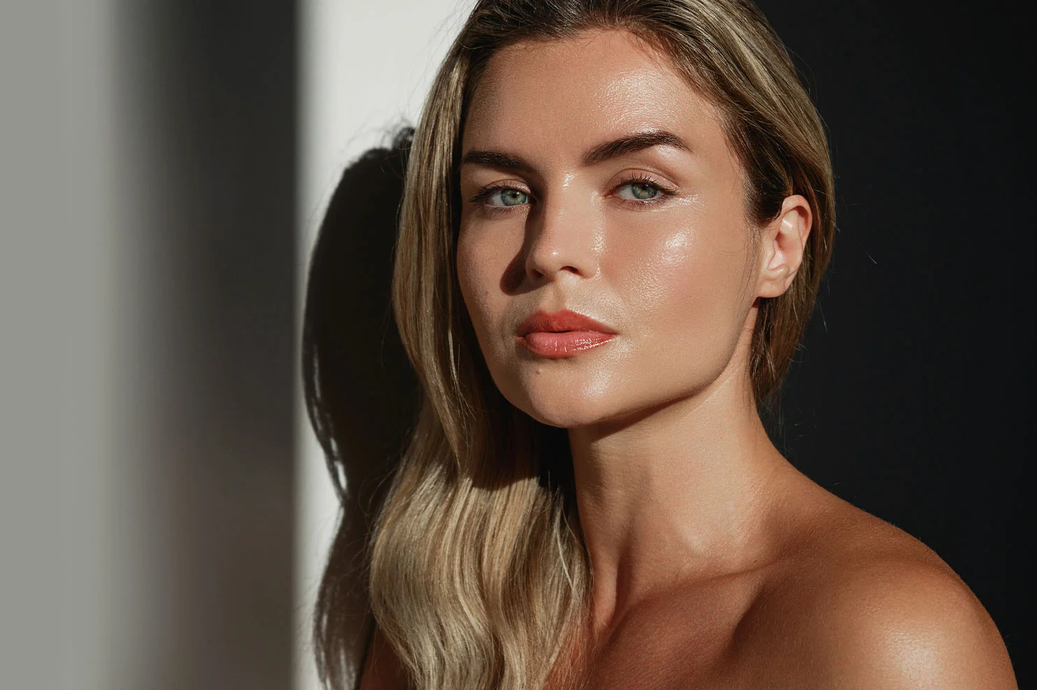 A close-up of a woman with blonde hair looking forward. High-contrast lighting creates a dramatic shadow on the wall behind her, highlighting the symmetrical contours and radiant finish of her skin.