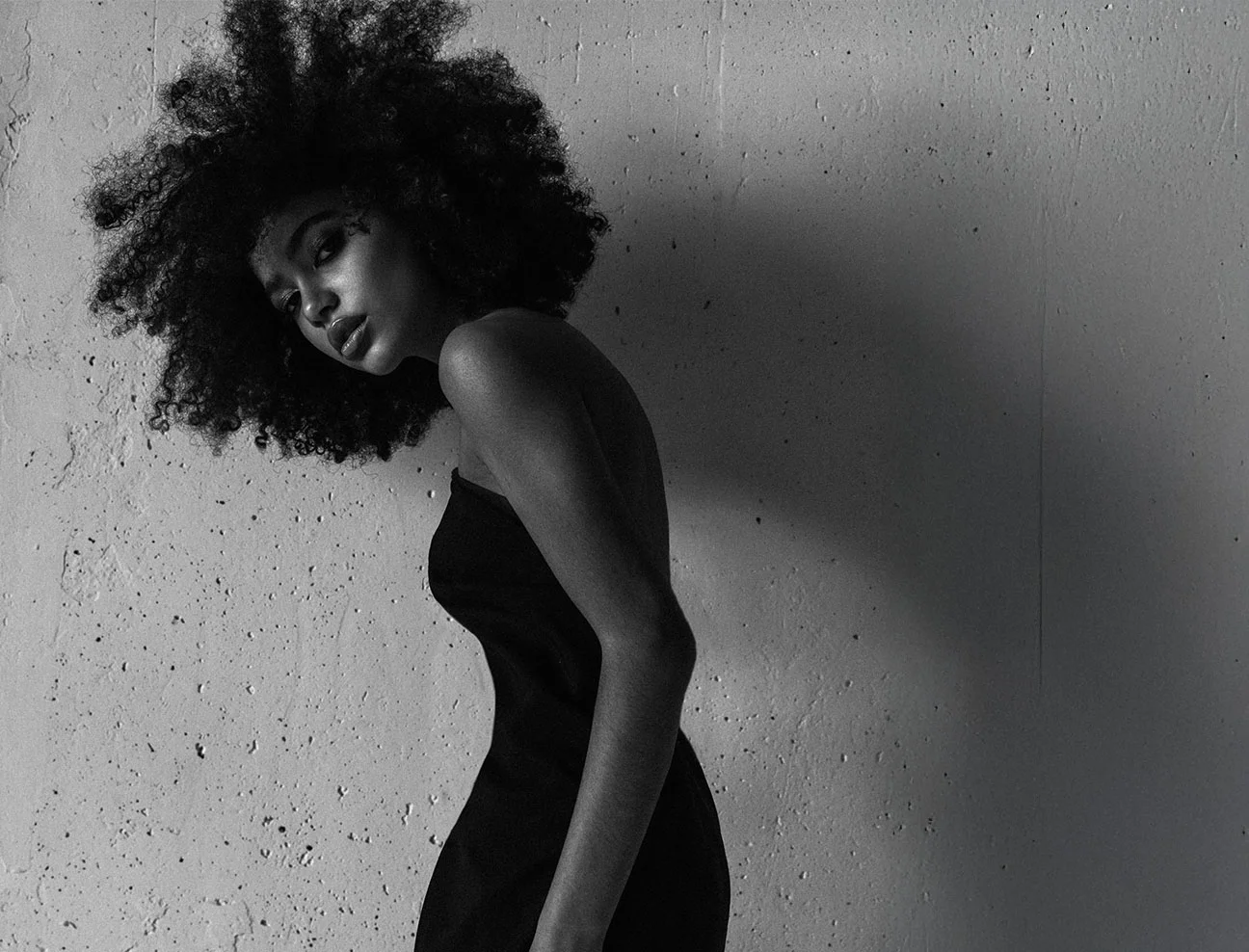 A black and white artistic shot of a woman with a large, voluminous afro. She is leaning forward against a textured concrete wall, looking back over her shoulder at the camera.