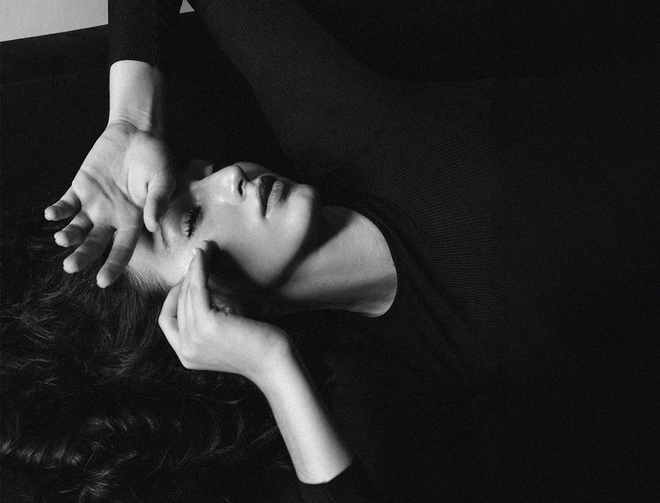 A black and white overhead shot of a woman with dark curly hair lying down. She has her hands raised near her forehead and eyes, creating a cinematic and relaxed aesthetic.