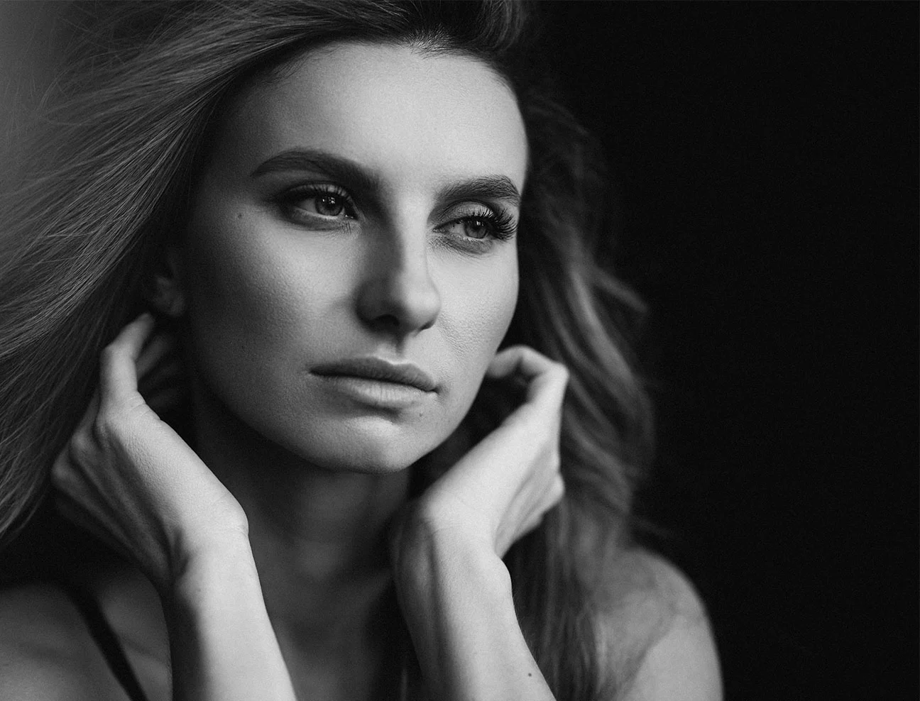 A black and white portrait of a woman looking off to the side with a pensive expression. She has her hands raised toward her face, softly touching her jawline, with her hair flowing slightly.