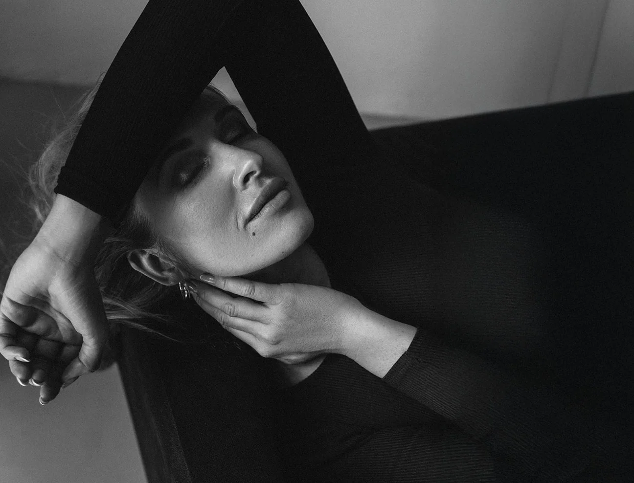 A black and white high-angle shot of a woman lying down with her eyes closed. One arm is bent over her head while her other hand gently touches her neck and jawline.