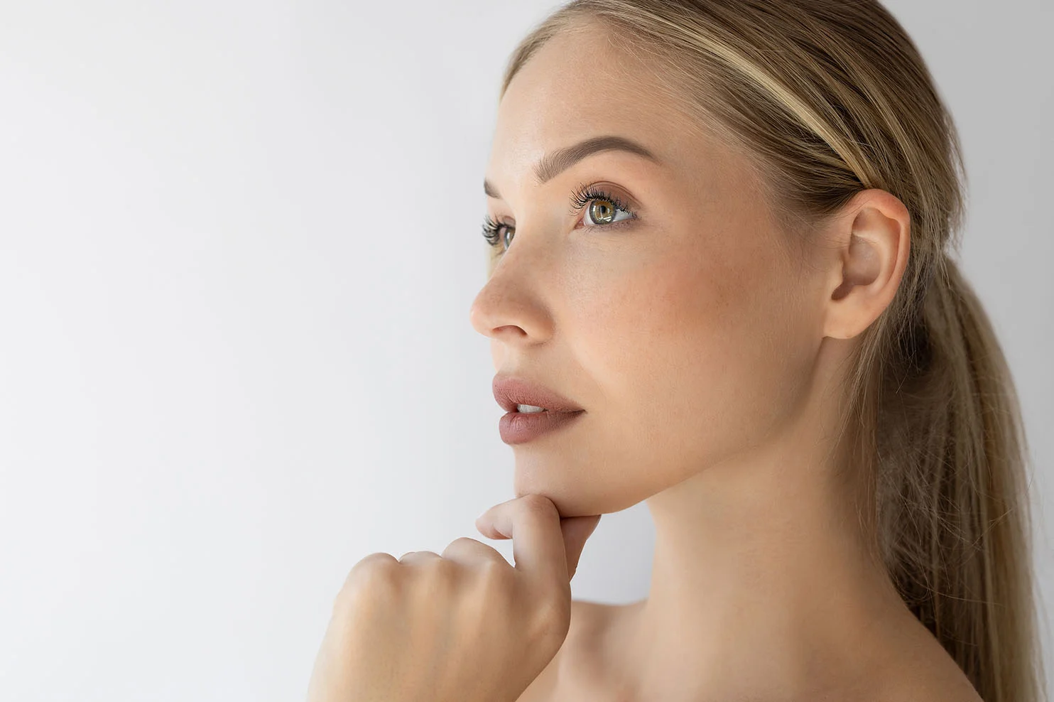 A side profile of a woman with a blonde ponytail, looking upward with her hand resting gently under her chin to highlight a defined jawline and neck area.