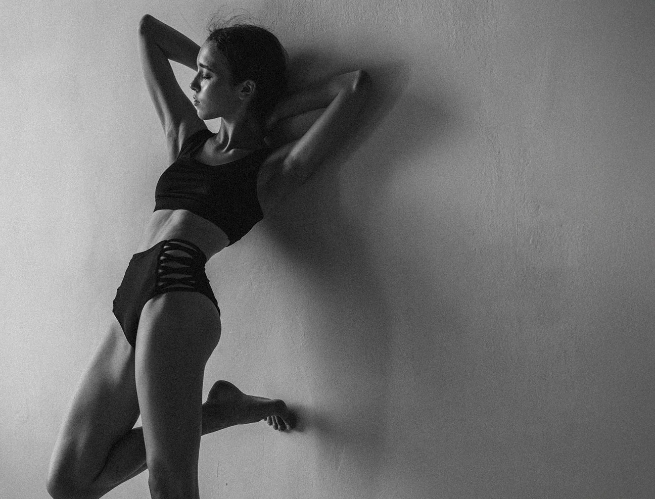 A black and white full-body silhouette of a woman in a black two-piece athletic set leaning against a textured wall. One leg is bent at the knee, and her arms are raised behind her head.