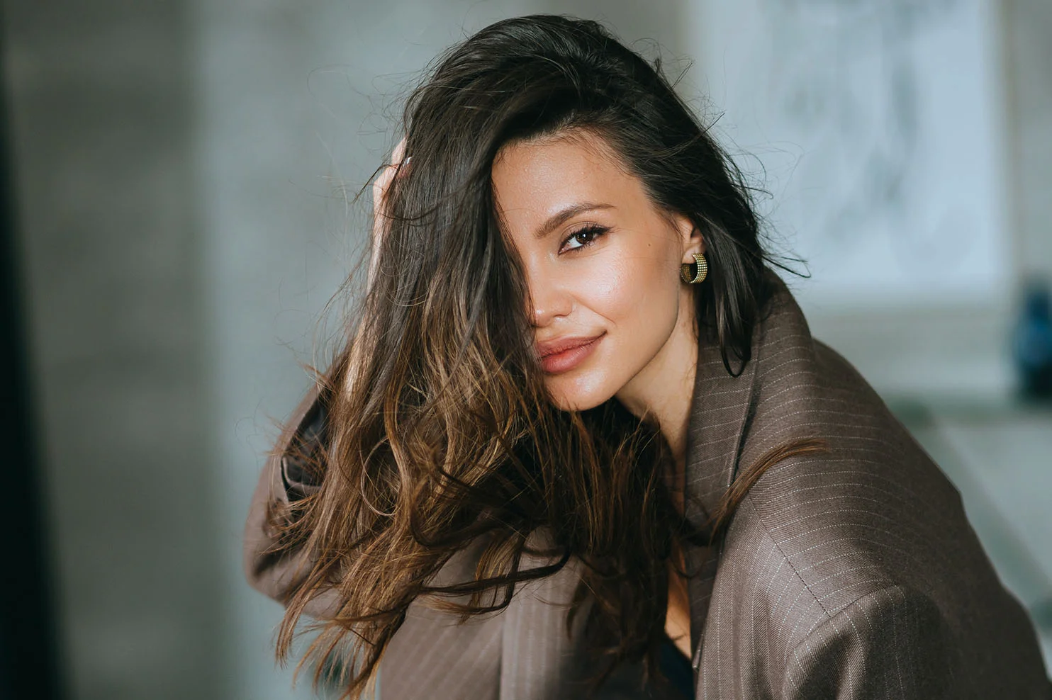 A woman with long, dark brown hair and a pinstripe blazer looking through her hair at the camera. The focus is on her full, natural-looking lips and polished aesthetic.