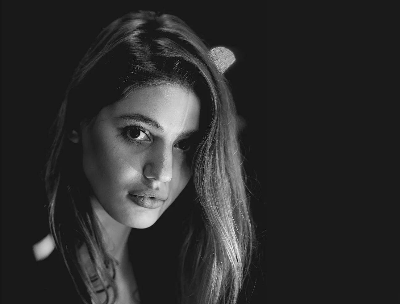 A black and white close-up of a woman's face, partially illuminated with soft light. Her hair falls naturally around her face as she looks directly into the lens.