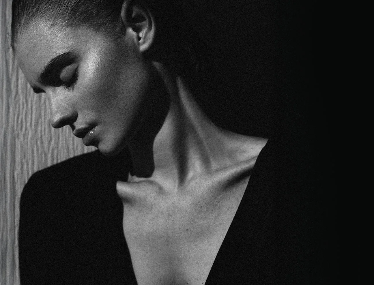 A high-contrast black and white artistic profile of a woman looking down. The lighting emphasizes the sharp lines of her jaw, neck, and collarbone against a dark background.