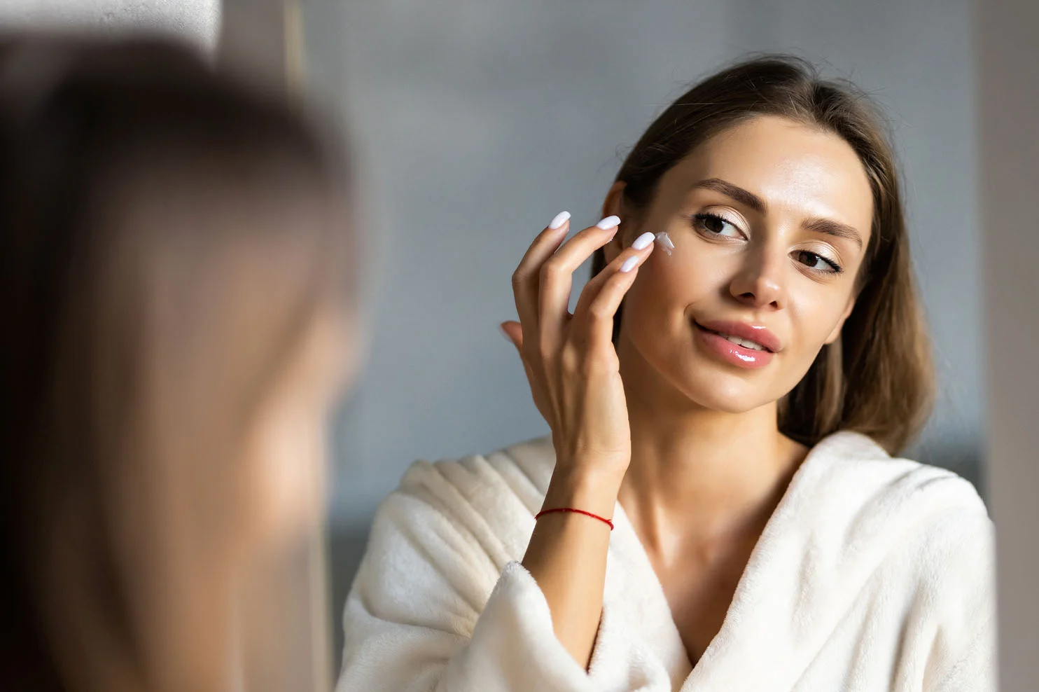 A woman in a white bathrobe stands before a bathroom mirror, carefully applying facial cream to her cheek. She has a gentle smile, looking at her reflection.