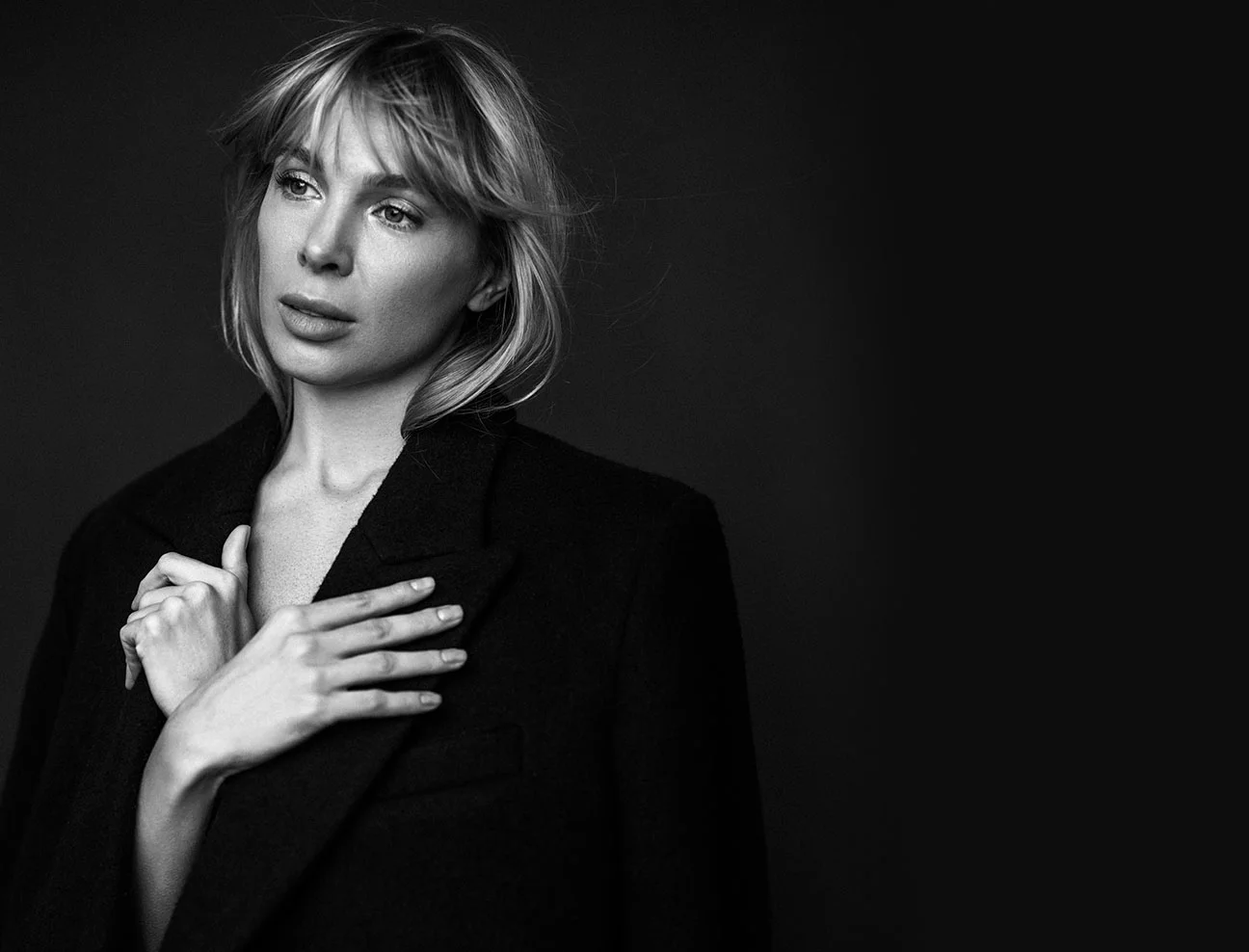 A black and white portrait of a woman with a blonde fringe and wavy hair. She is wearing a dark wool coat and has her hands clasped near her chest, looking away with a contemplative gaze.