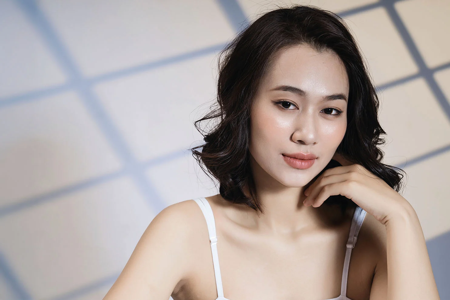 A woman with short, dark wavy hair and a white camisole poses against a background with a geometric window-pane shadow. She has a serene expression and glowing, even skin.