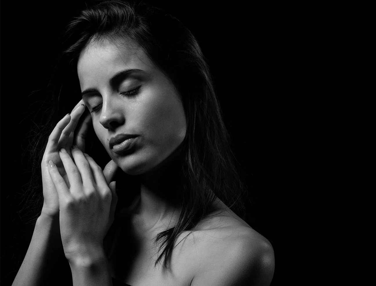 A black and white image of a woman with her eyes closed and hands gently touching her temples and jaw. The shot is set against a dark background, highlighting her facial features.