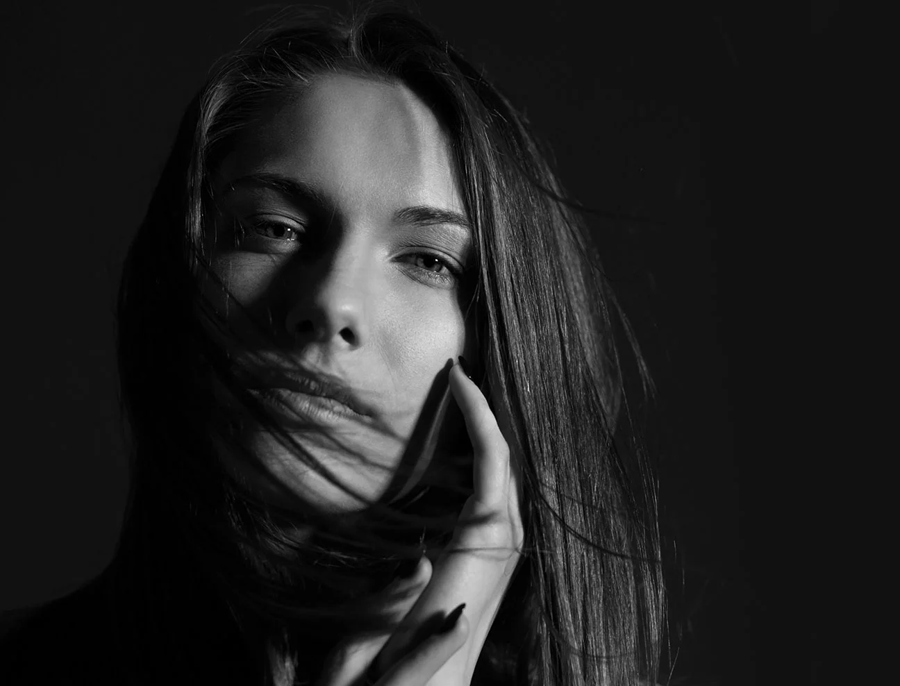 A black and white portrait of a woman with long, straight hair blowing across her face. She has her hand resting against her cheek and is looking directly at the camera with a piercing gaze.