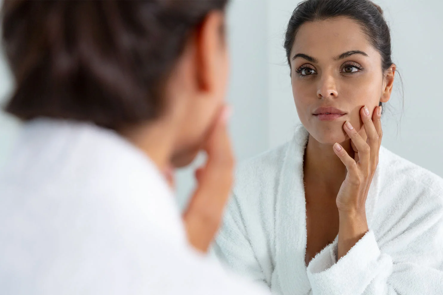 A woman in a white plush bathrobe looks into a mirror, gently touching her cheek. Her reflection shows a focused expression and a healthy, rejuvenated facial glow.