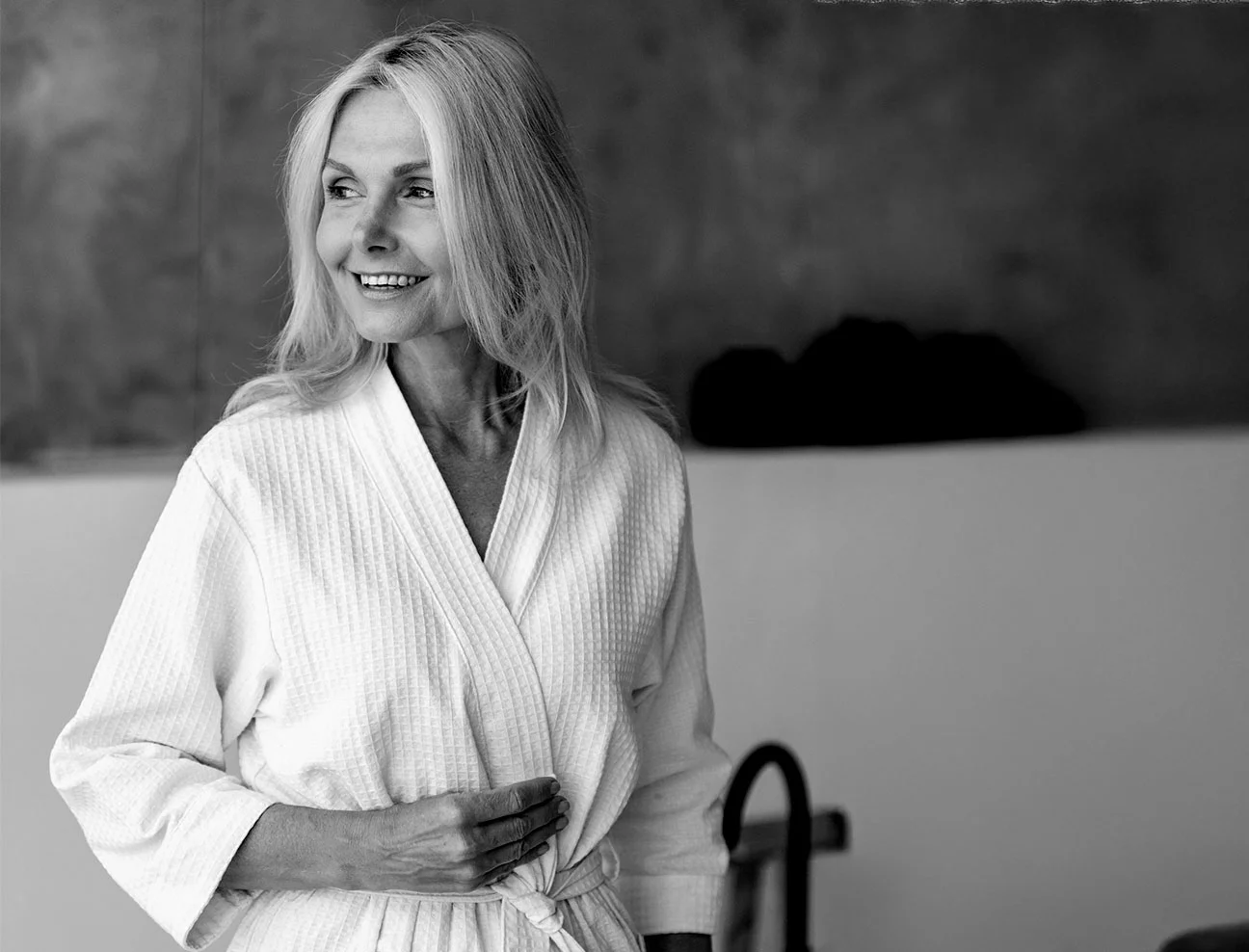 A black and white image of a mature woman with long, light hair wearing a white waffle-weave spa robe. She is smiling and looking off-camera, conveying a sense of health and vitality.