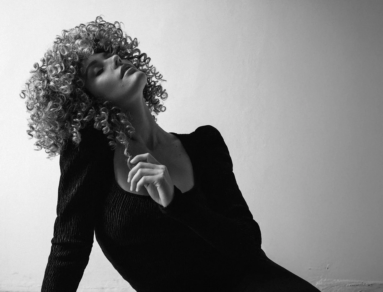 A black and white artistic shot of a woman with voluminous, curly hair. She is sitting on the floor, leaning back with her eyes closed and chin tilted upward in a pose of relaxation.