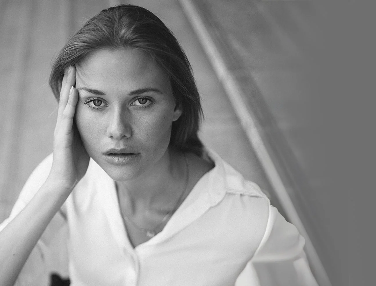 A black and white, soft-focus photo of a woman in a white button-up shirt looking upward with a serene expression. She has one hand resting against her temple.