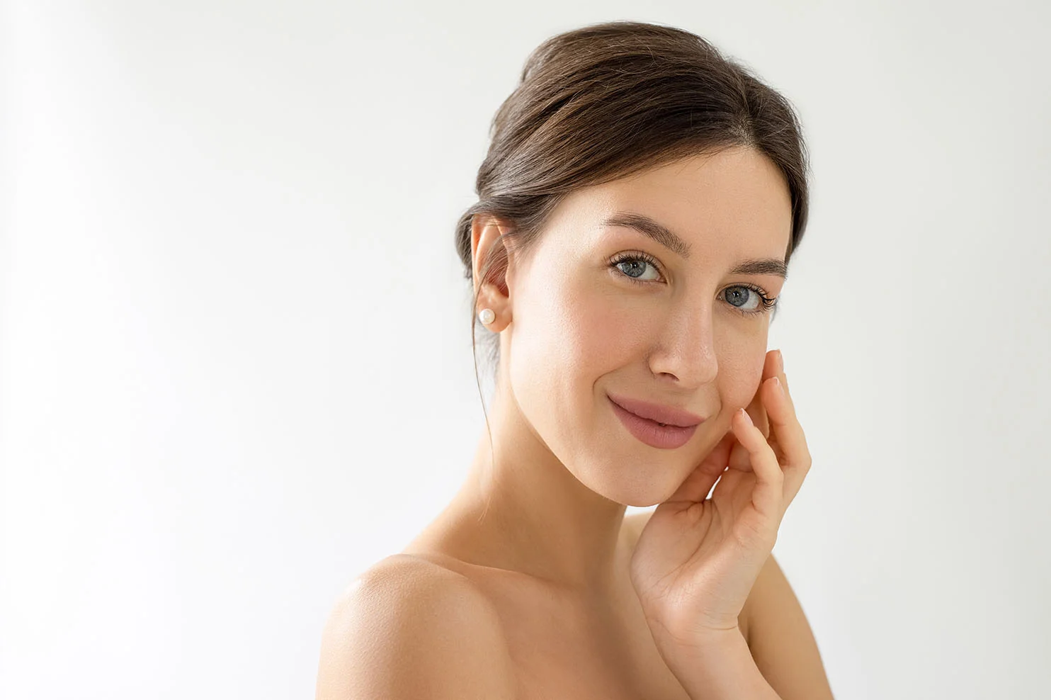 A woman with her hair in a sleek updo and pearl earrings smiles warmly. She gently touches her cheek, emphasizing a healthy, hydrated glow against a bright white background.