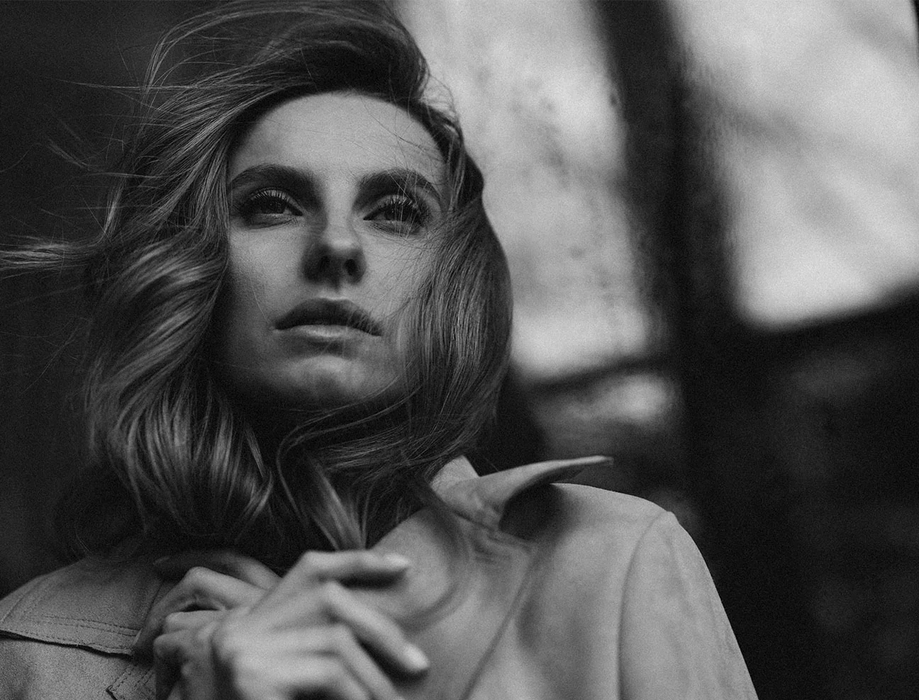 A black and white, slightly grainy close-up of a woman looking upward with an expectant expression. Her hair is blowing in the wind, and she is holding the collar of her light-colored coat.