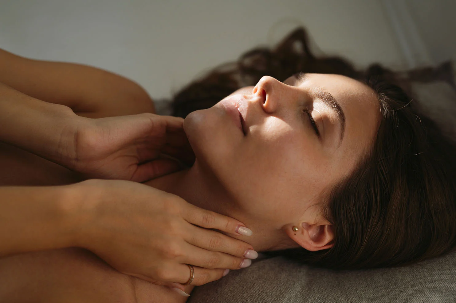 A close-up, top-down view of a woman lying down with her eyes closed in a state of deep relaxation. Two hands are shown gently supporting her neck and jawline, with warm sunlight creating soft patterns across her face.