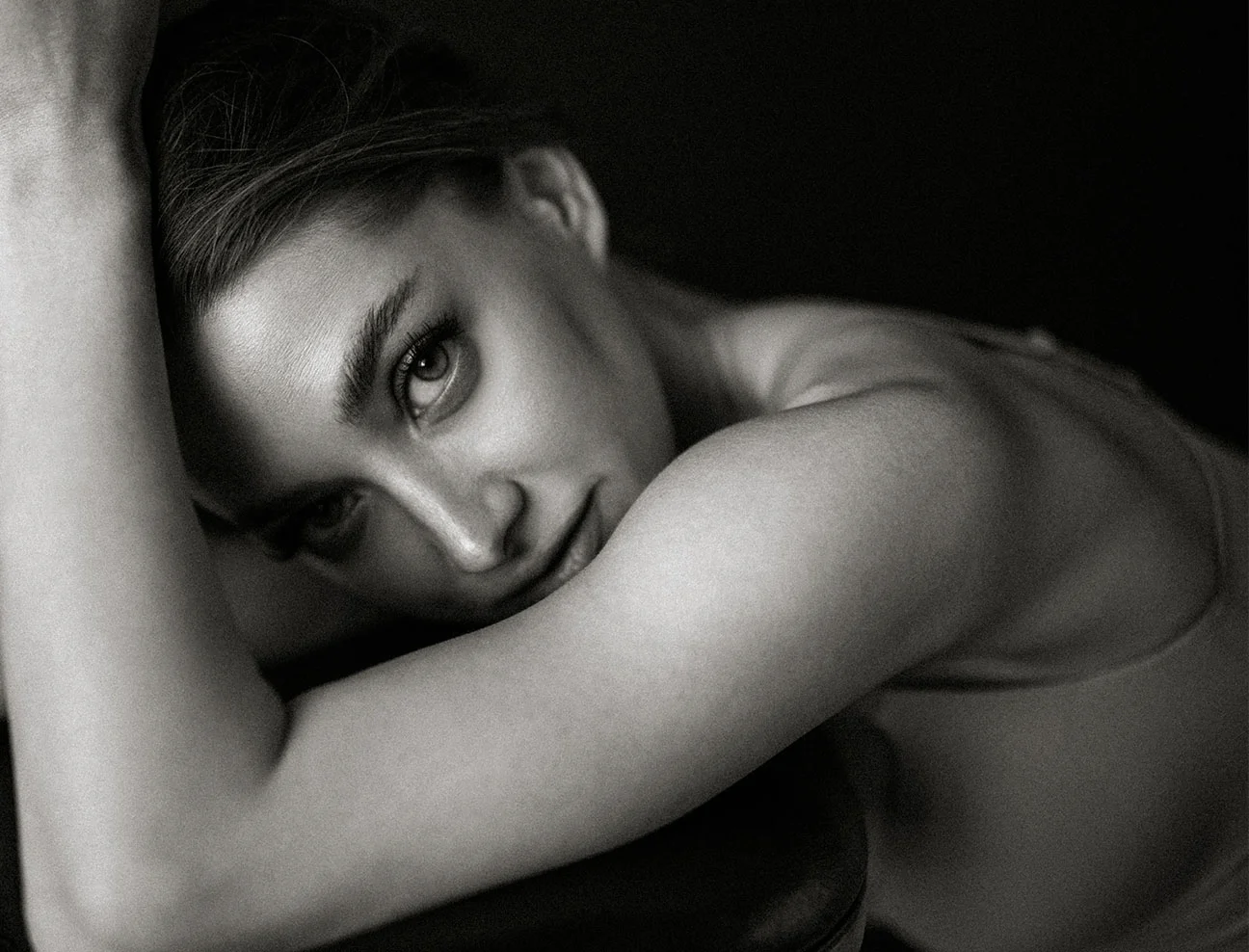 A black and white, close-up shot of a woman leaning forward and looking into the camera with a soft, natural expression. Her arm is raised, framing the side of her face.