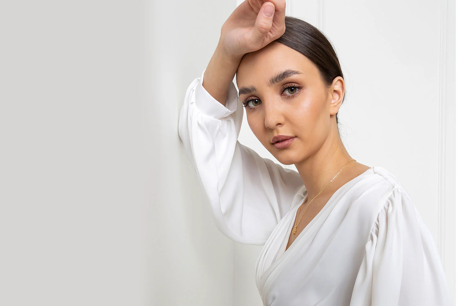 A woman in a white silk blouse leans her head against a white wall. She looks directly at the camera with a neutral, sophisticated expression, showcasing even skin tone.
