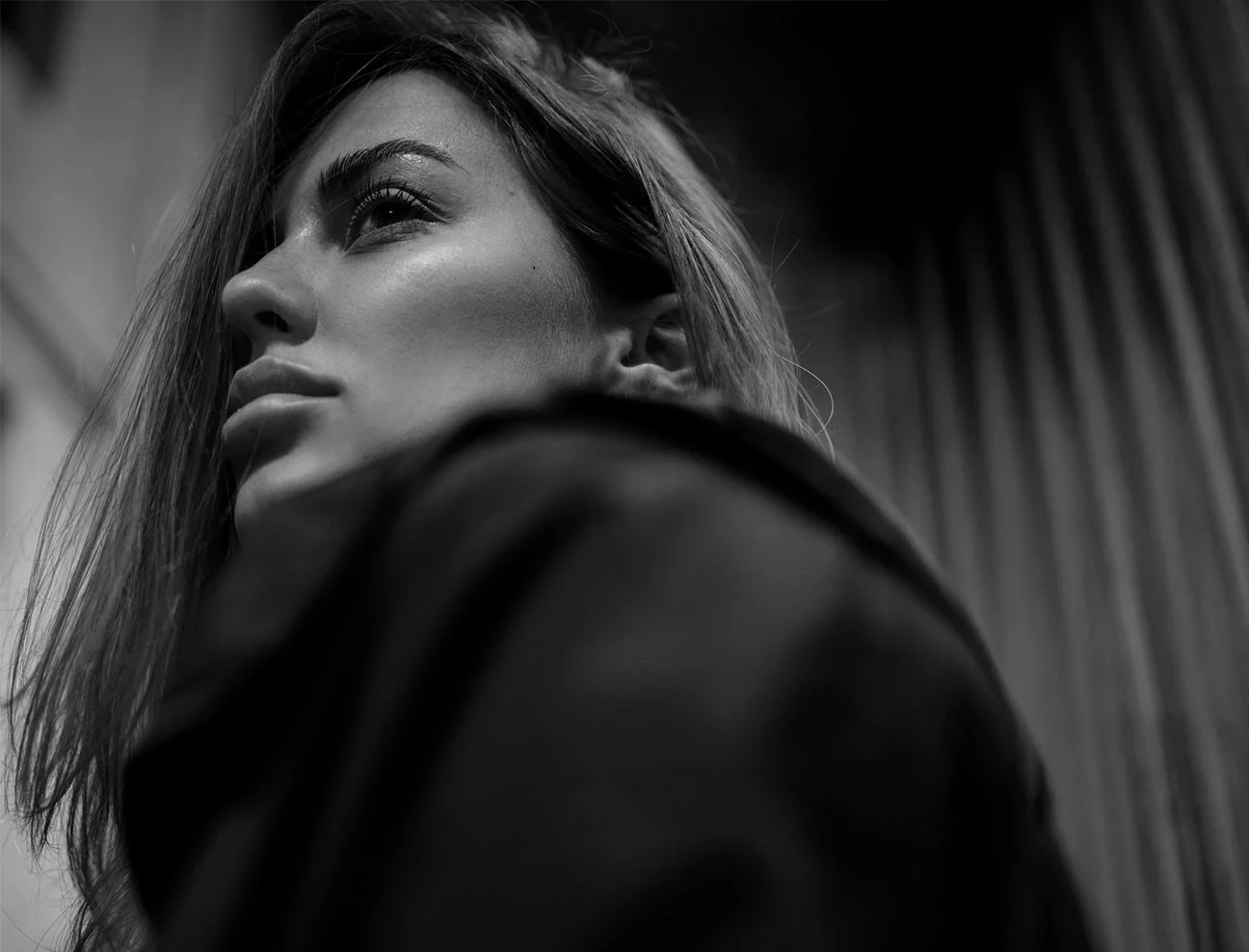 A black and white, low-angle profile of a woman looking upward toward a light source. The lighting emphasizes her facial features and jawline against a soft, blurred background.