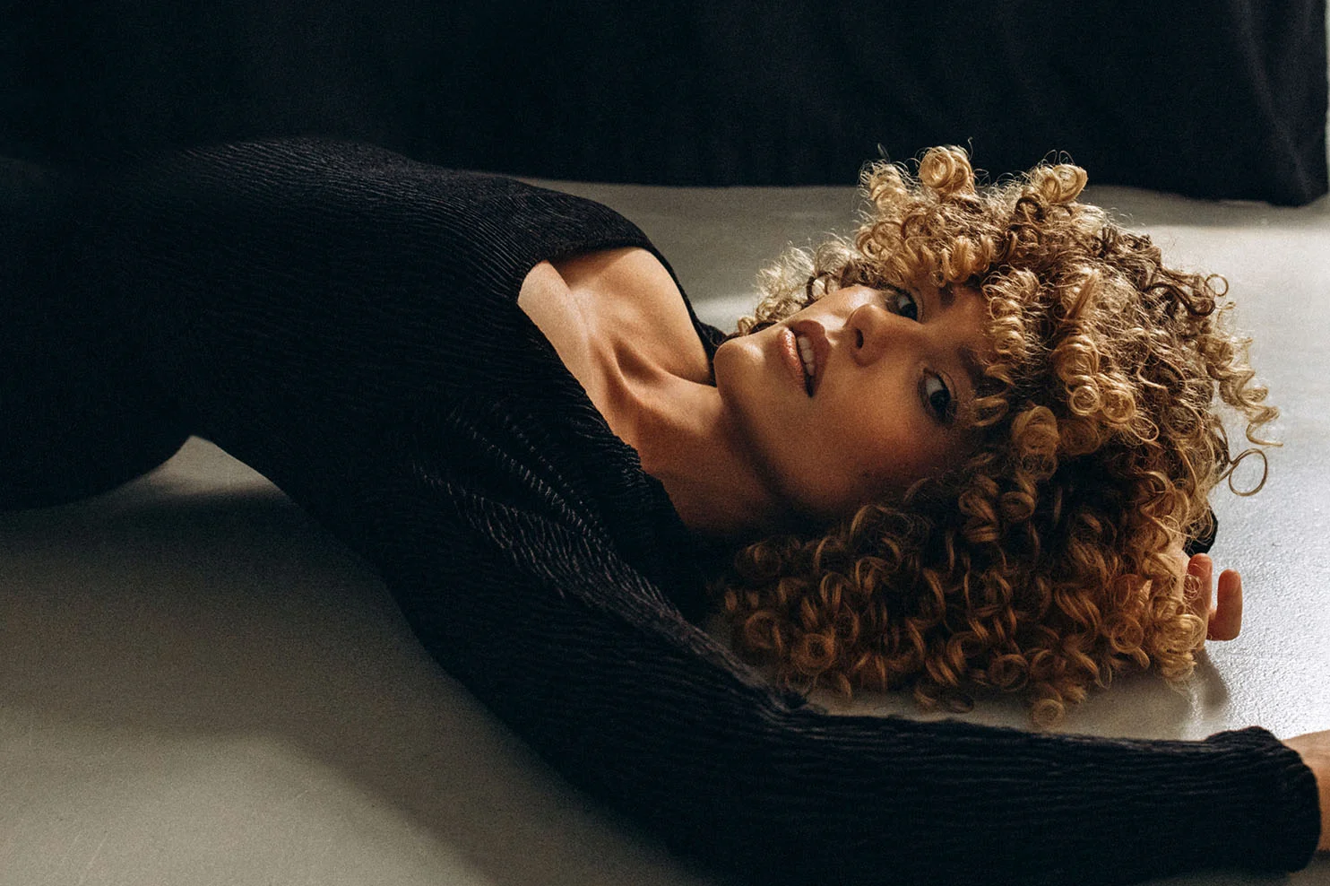 An artistic, moody shot of a woman with voluminous blonde curls lying on a grey floor. She is wearing a black top and looking upward, with the lighting highlighting the contours of her face.