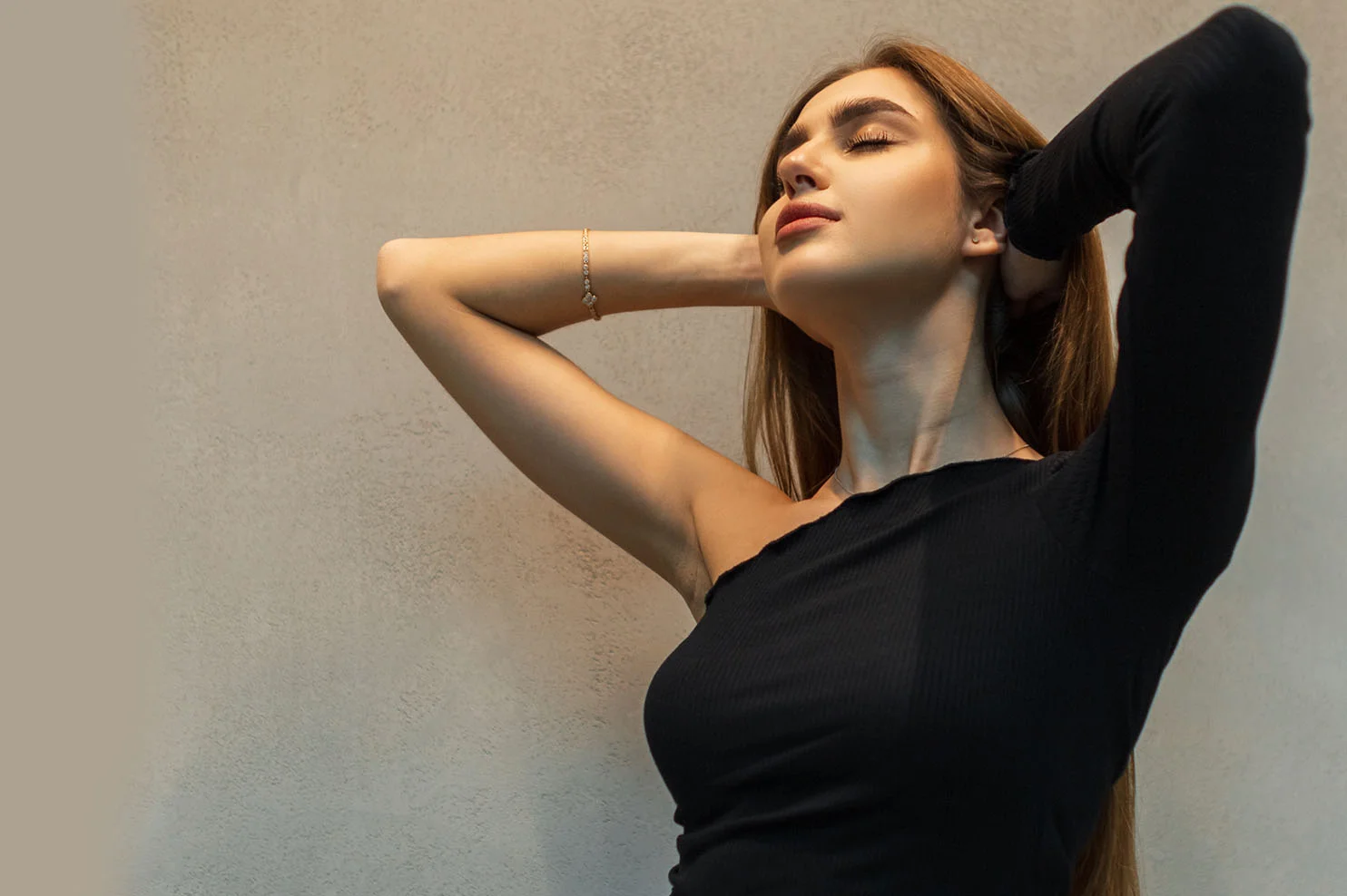 A woman in a black one-shoulder top tilts her head back with her eyes closed and hands behind her head. The lighting emphasizes the smooth, refreshed skin of her face and neck.